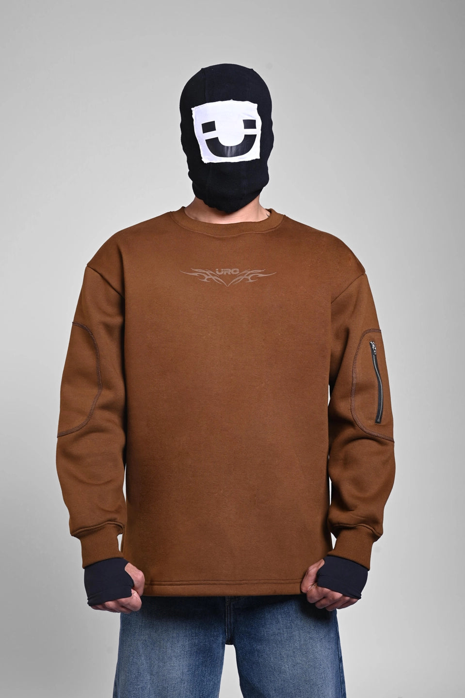Person in a brown sweatshirt with a small chest logo and a zippered pocket on the sleeve, wearing a black face mask.