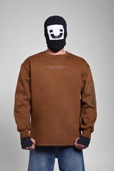 Person in a brown sweatshirt with a small chest logo and a zippered pocket on the sleeve, wearing a black face mask.