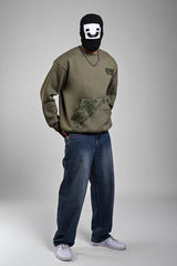 Person in an olive green graphic sweatshirt and blue jeans wearing a white-on-black graphic balaclava.