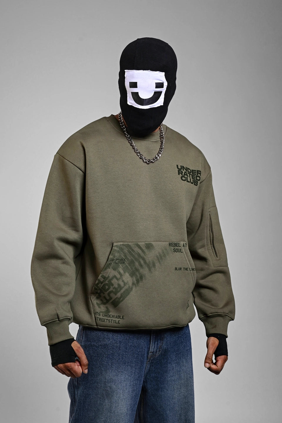 Person wearing an olive green "Underrated Club" sweatshirt with graphic text on the pocket and a silver chain.
