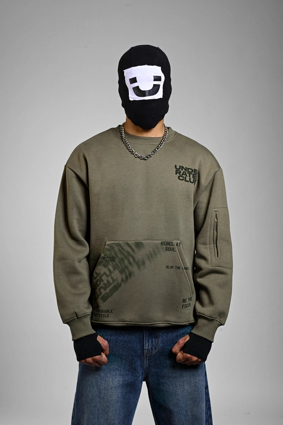 Person wearing an olive green "Underrated Club" sweatshirt with graphic text on the pocket and a silver chain.
