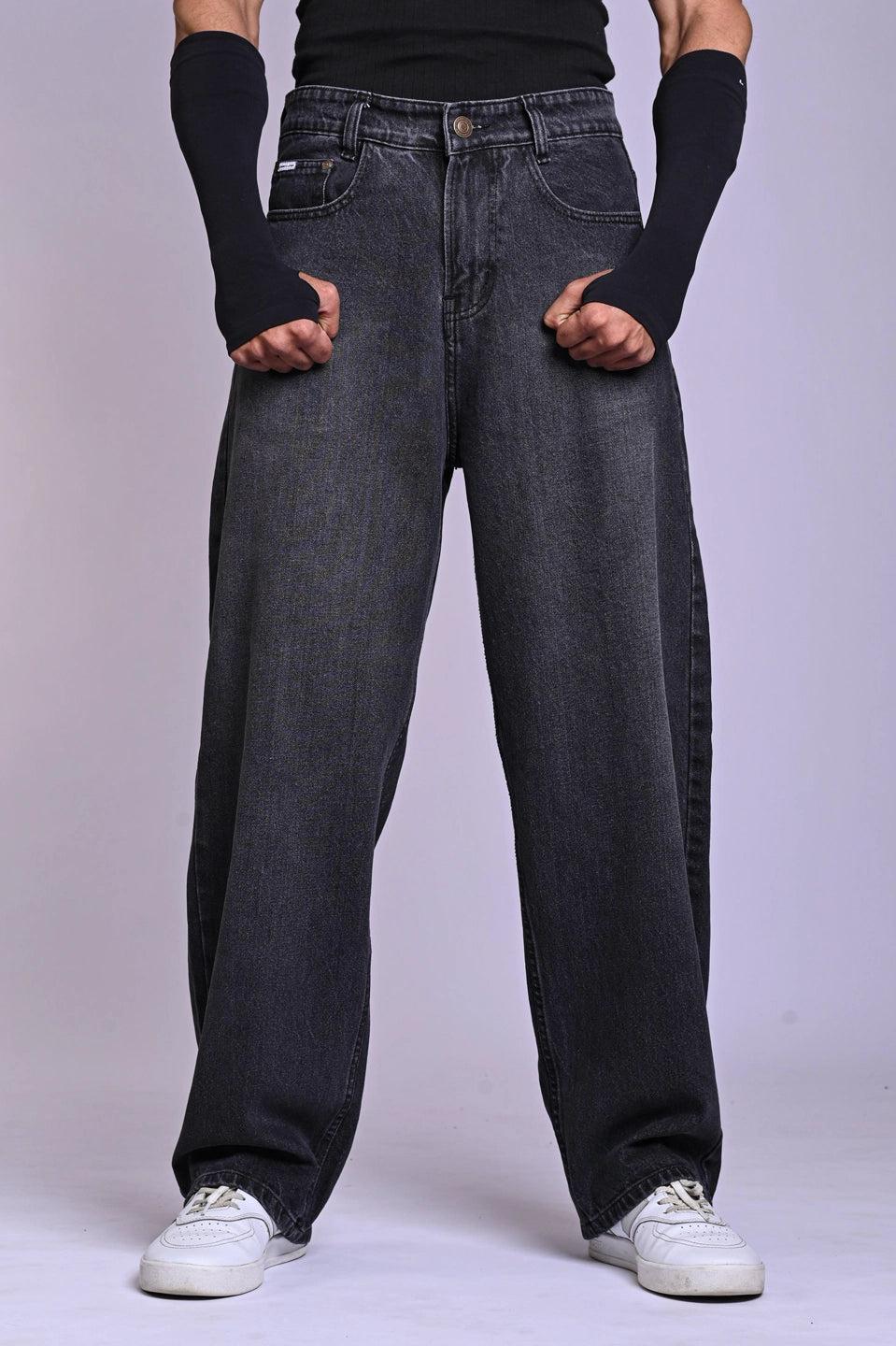 Front view of a person wearing oversized, dark grey acid-wash jeans and white sneakers against a plain background.