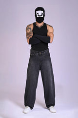 A tattooed man in a black tank top, arm sleeves, and a graphic balaclava wearing baggy charcoal acid-wash jeans.