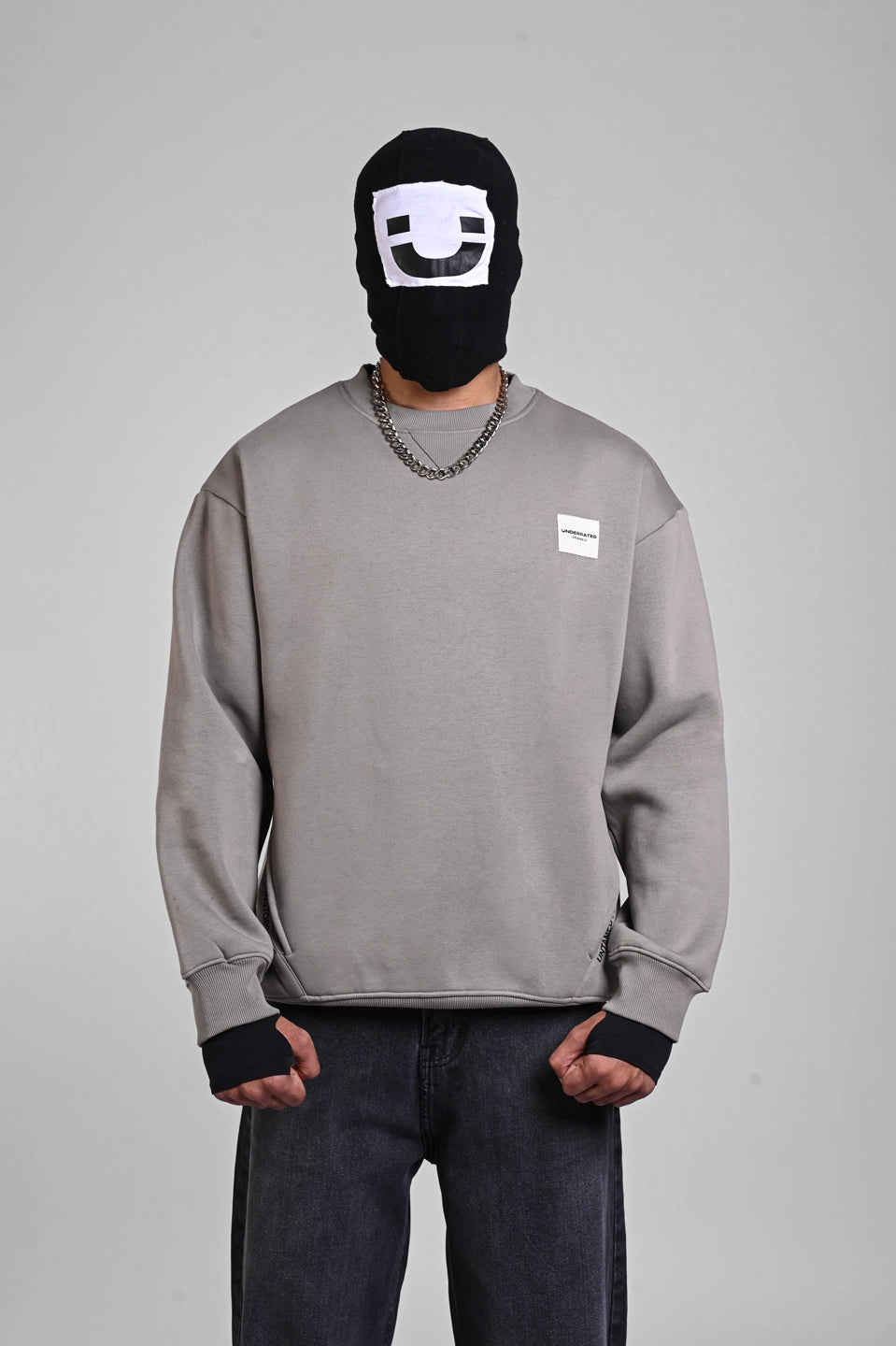 Front view of a person wearing a grey sweatshirt with a small white chest patch logo and a graphic balaclava.