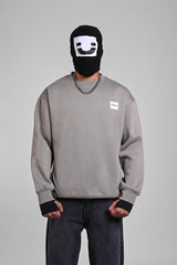 Front view of a person wearing a grey sweatshirt with a small white chest patch logo and a graphic balaclava.
