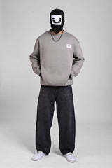 Full-body view of a person in a grey sweatshirt, dark baggy jeans, and white sneakers, wearing a black face mask.