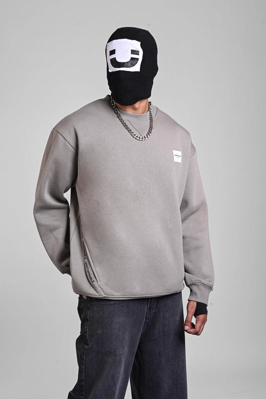 Full-body view of a person in a grey sweatshirt, dark baggy jeans, and white sneakers, wearing a black face mask.