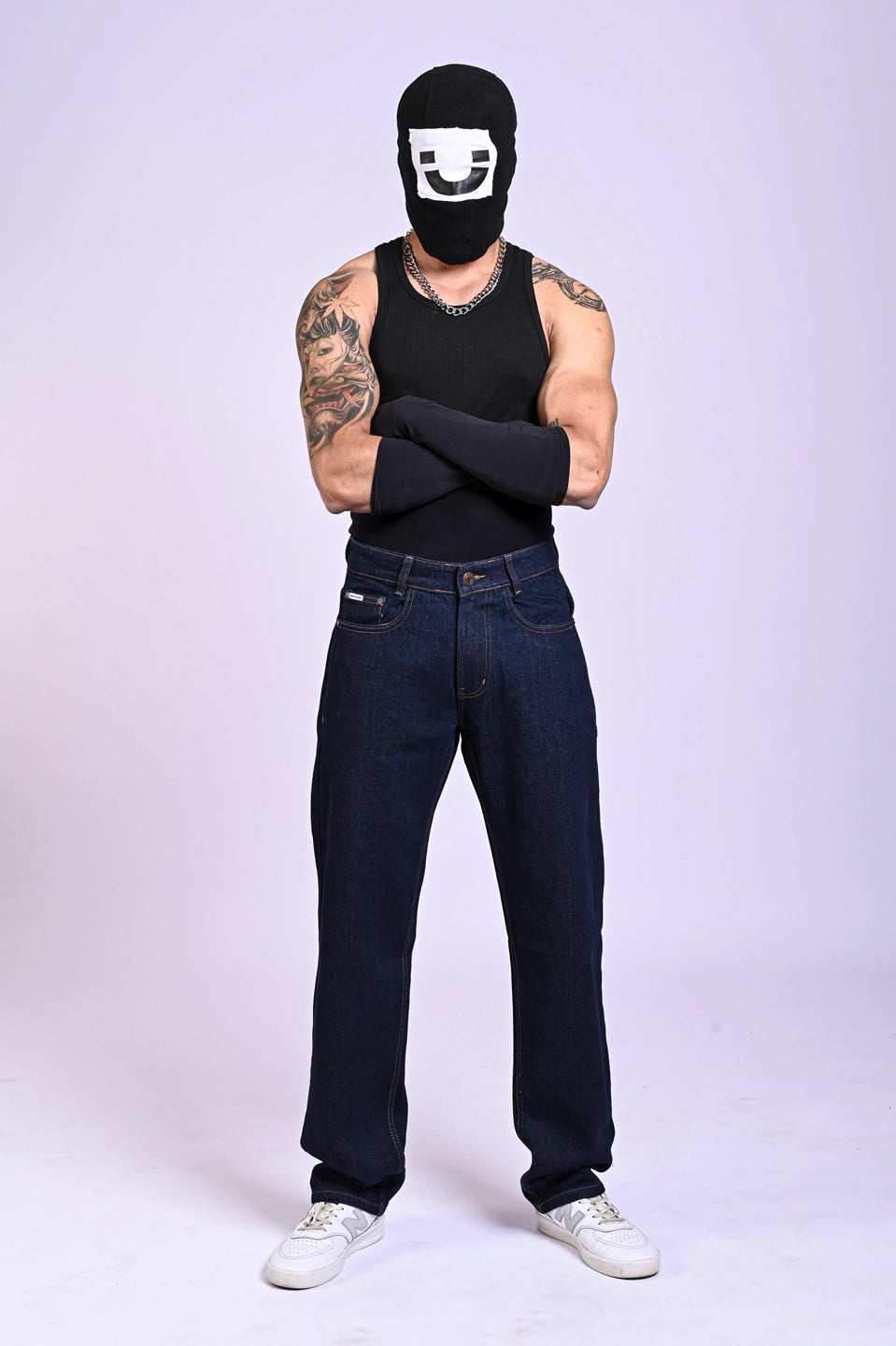 Person in a black tank top, balaclava, and arm sleeves posing in baggy dark indigo blue jeans.