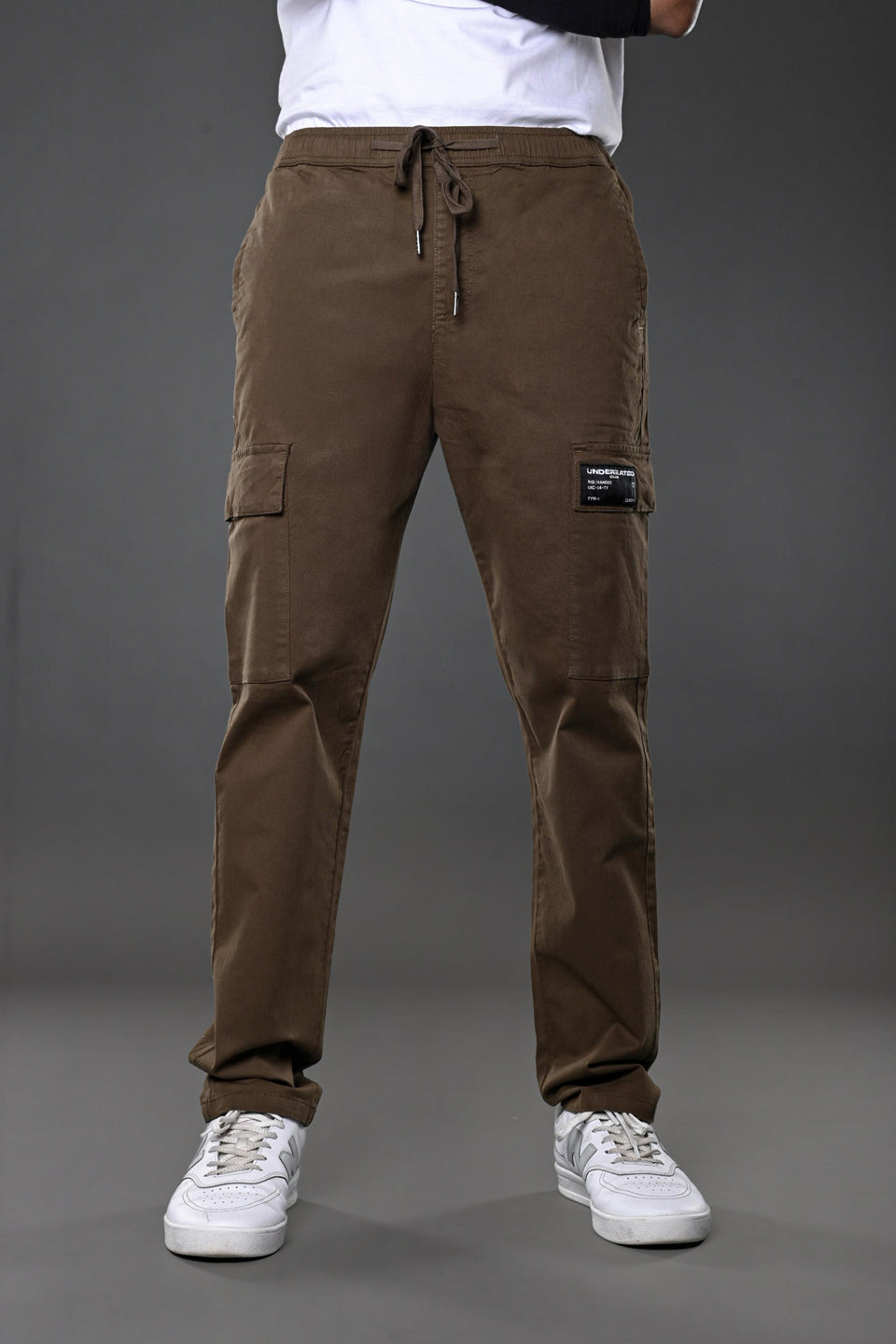 Man modeling brown UNDERRATED CLUB drawstring cargo pants with branded patch, flap pockets, white t-shirt, black wristband, and white sneakers against grey background.
