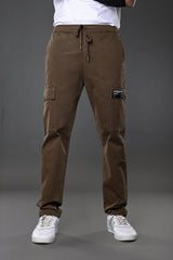 Man modeling brown UNDERRATED CLUB drawstring cargo pants with branded patch, flap pockets, white t-shirt, black wristband, and white sneakers against grey background.
