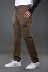 Man modeling brown UNDERRATED CLUB drawstring cargo pants with branded flap pocket, white t-shirt, black wristband, and white Nike sneakers against grey background.
