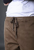Close-up of brown UNDERRATED CLUB drawstring cargo pants waistband detail on model, showing adjustable drawstrings, white t-shirt edge, and black sleeve against grey background.