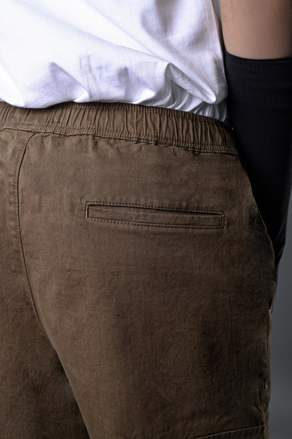Back view of brown UNDERRATED CLUB drawstring cargo pants on model, showing rear pockets, elastic waistband, white t-shirt, and black wristbands against grey background.