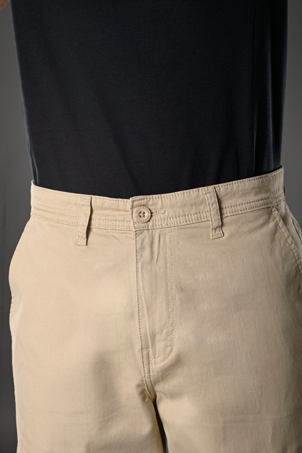 Close-up of beige UNDERRATED cargo pants waistband on model wearing black t-shirt, featuring button closure and drawstring details against grey background.
