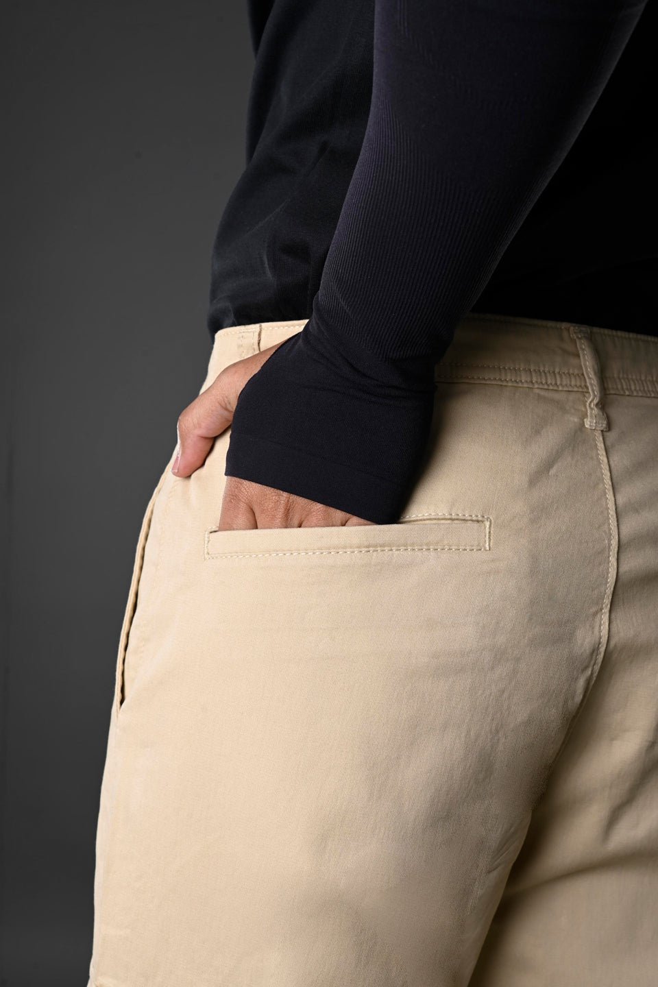 Back view of beige UNDERRATED cargo pants on model in black long-sleeve top, hand in rear pocket, against grey background.
