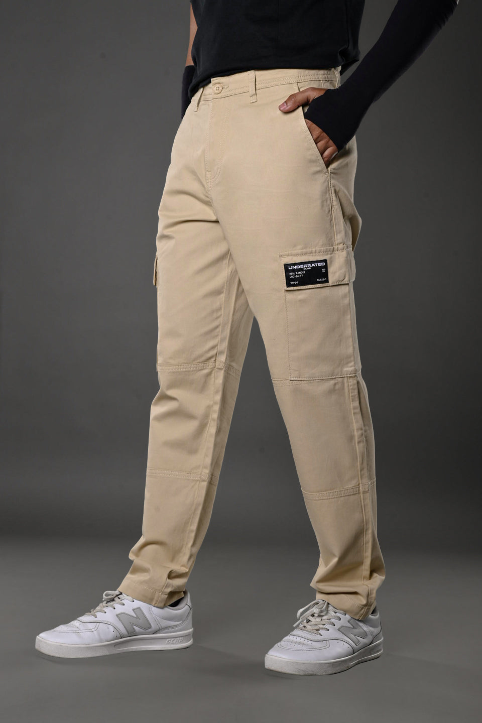 Full front view of man in beige UNDERRATED cargo pants, black top, wristbands, white sneakers posing confidently.
