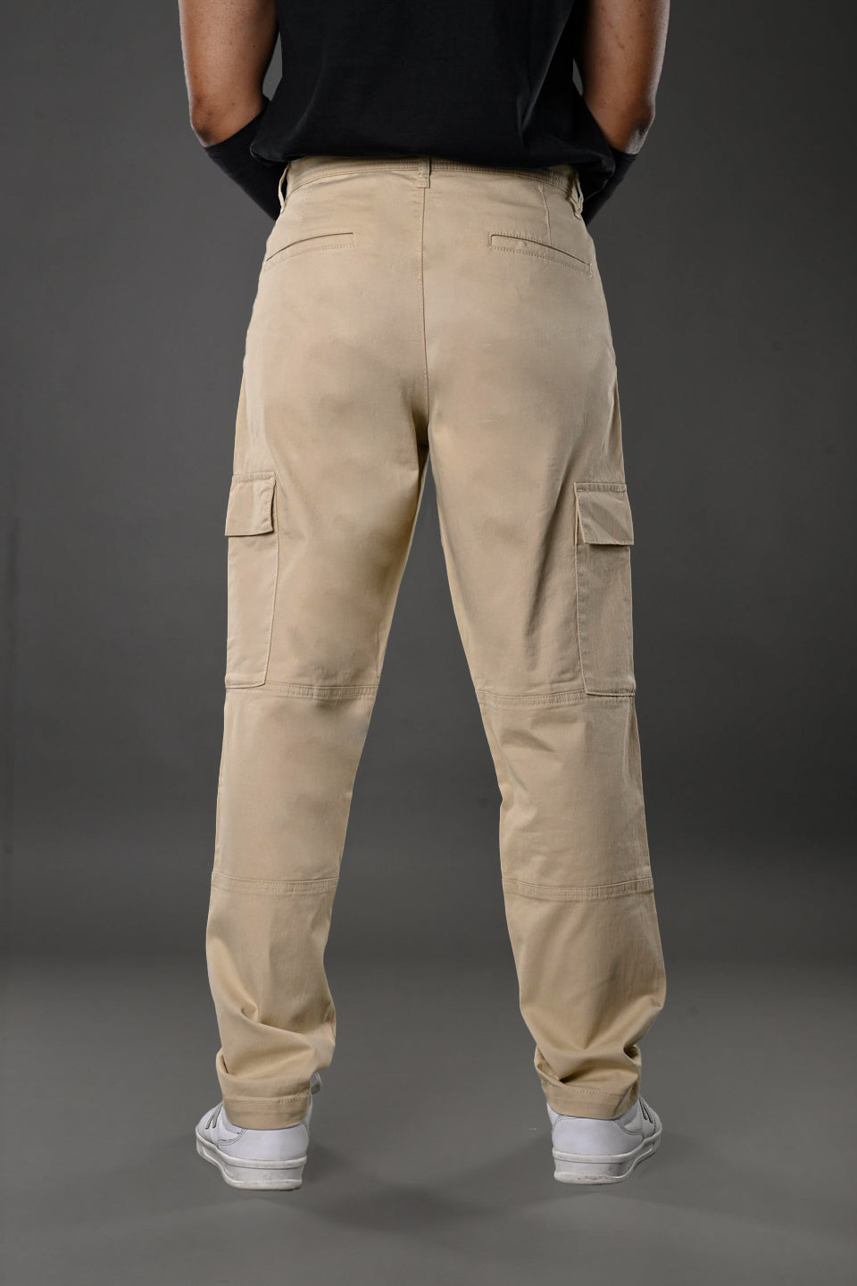 Back view of man in beige UNDERRATED cargo pants with wristbands, black t-shirt, and white sneakers against grey background.
