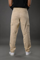 Back view of man in beige UNDERRATED cargo pants with wristbands, black t-shirt, and white sneakers against grey background.
