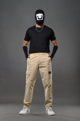 Man wearing black smiley mask, black t-shirt with chain necklace, wristbands, beige UNDERRATED cargo pants, and white sneakers posing confidently against grey background.