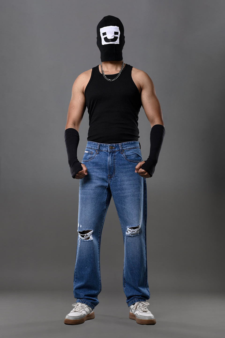 Full-body man wearing white smiley balaclava black tank top slim blue jeans white sneakers gray background.
