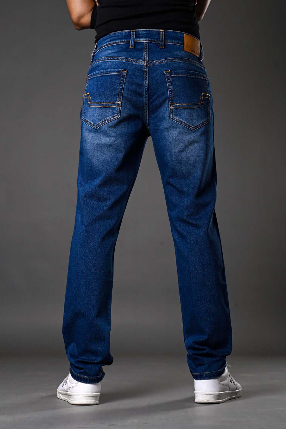Full rear view man wearing blue distressed denim jeans white sneakers black top, showcasing back pockets stitching.
