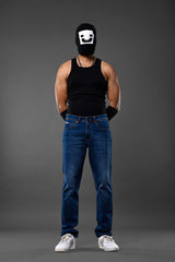 Full-body man wearing white smiley balaclava black tank top slim blue jeans white sneakers gray background.

