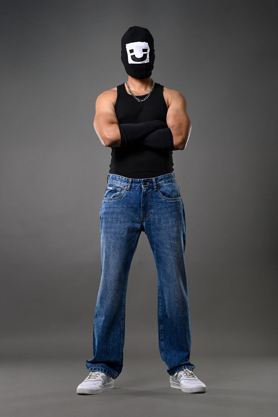 Full-body man wearing white smiley balaclava black tank top slim blue jeans white sneakers gray background.
