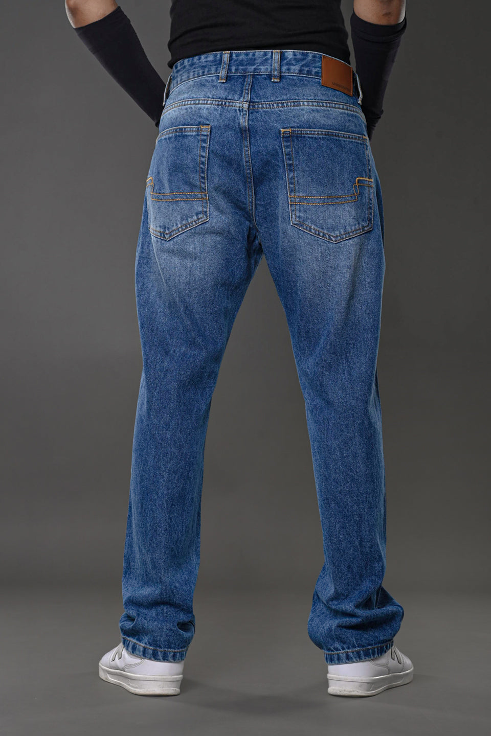 Rear full view man in light blue relaxed fit jeans black top arm cuffs white sneakers gray background Underrated Club denim.
