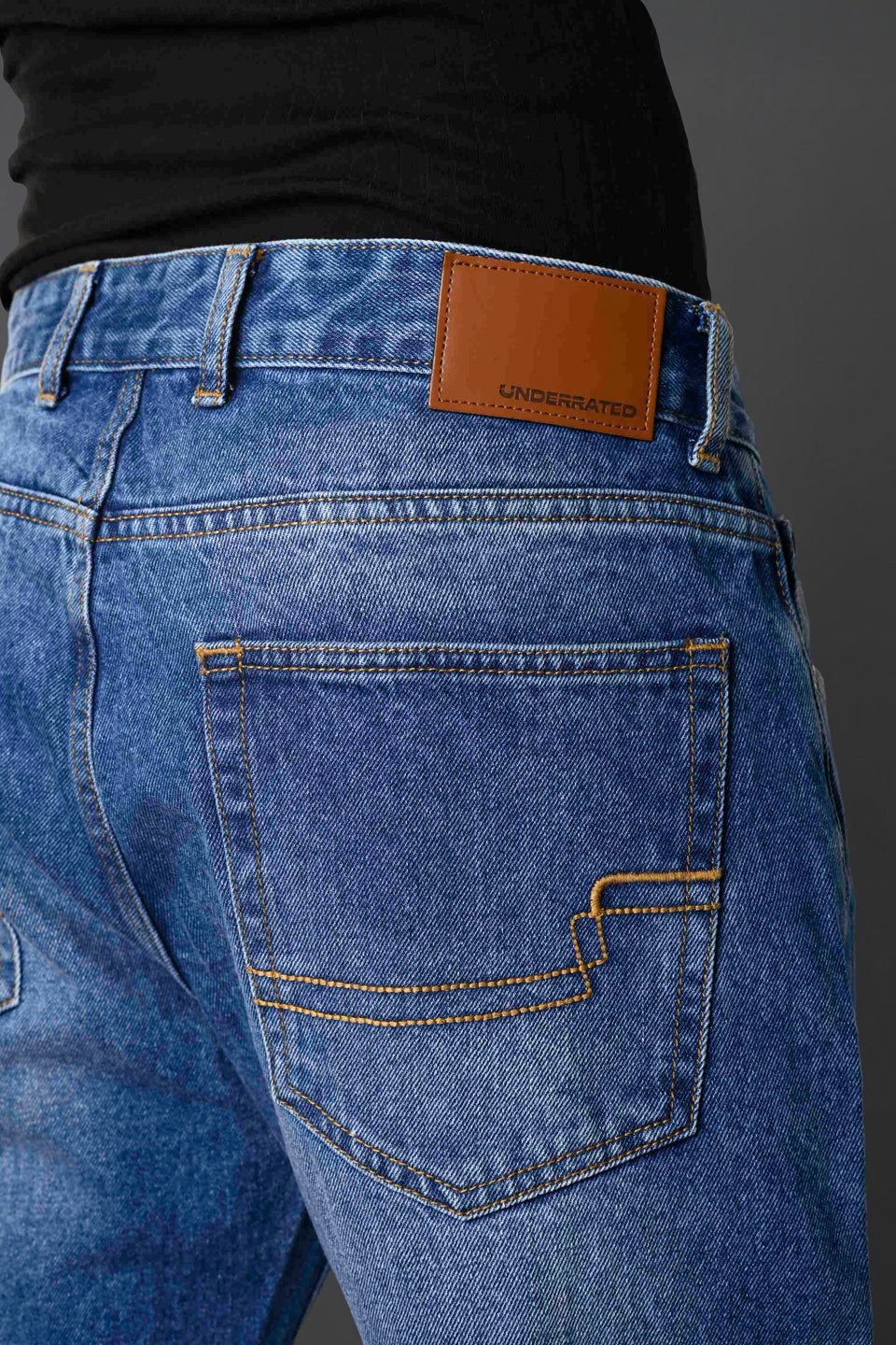 Close-up back pocket tan leather Underrated Club label yellow zigzag stitching light blue jeans detail.
