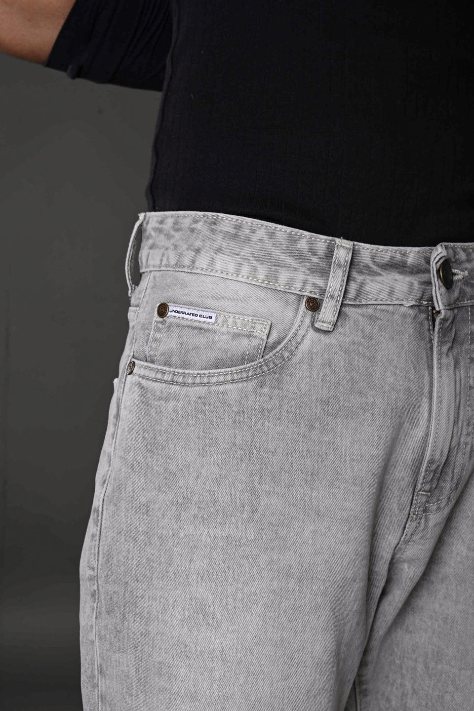 Underrated Club baggy jeans front pocket brass rivets light gray wash closeup branded apparel.
