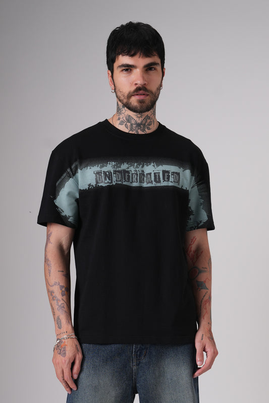 Man wearing a black Underrated graphic t-shirt and blue jeans standing against a plain grey background, showing tattoos on his arms and neck.