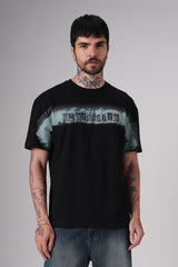 Man wearing a black Underrated graphic t-shirt and blue jeans standing against a plain grey background, showing tattoos on his arms and neck.