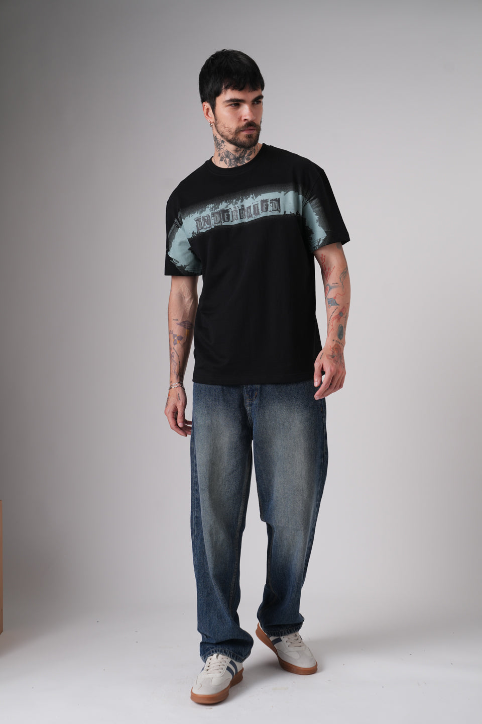 Full-body studio shot of a man wearing a black Underrated graphic t-shirt, relaxed blue jeans, and white sneakers against a plain grey background.