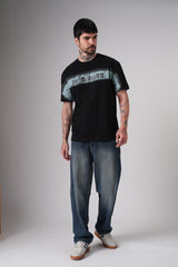 Full-body studio shot of a man wearing a black Underrated graphic t-shirt, relaxed blue jeans, and white sneakers against a plain grey background.