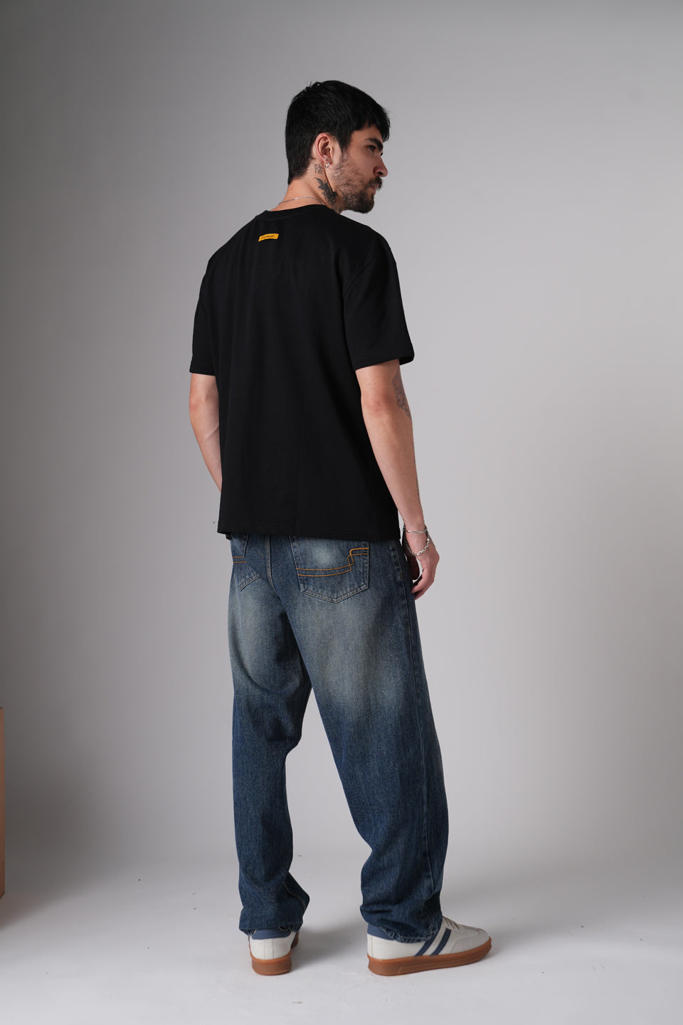 Three-quarter back full-body shot of the model in the black t-shirt and loose blue jeans, standing casually in a studio setting.