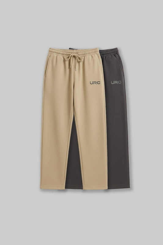 Flat lay of two pairs of relaxed-fit "URC" drawstring trousers in beige and charcoal grey against a grey background.