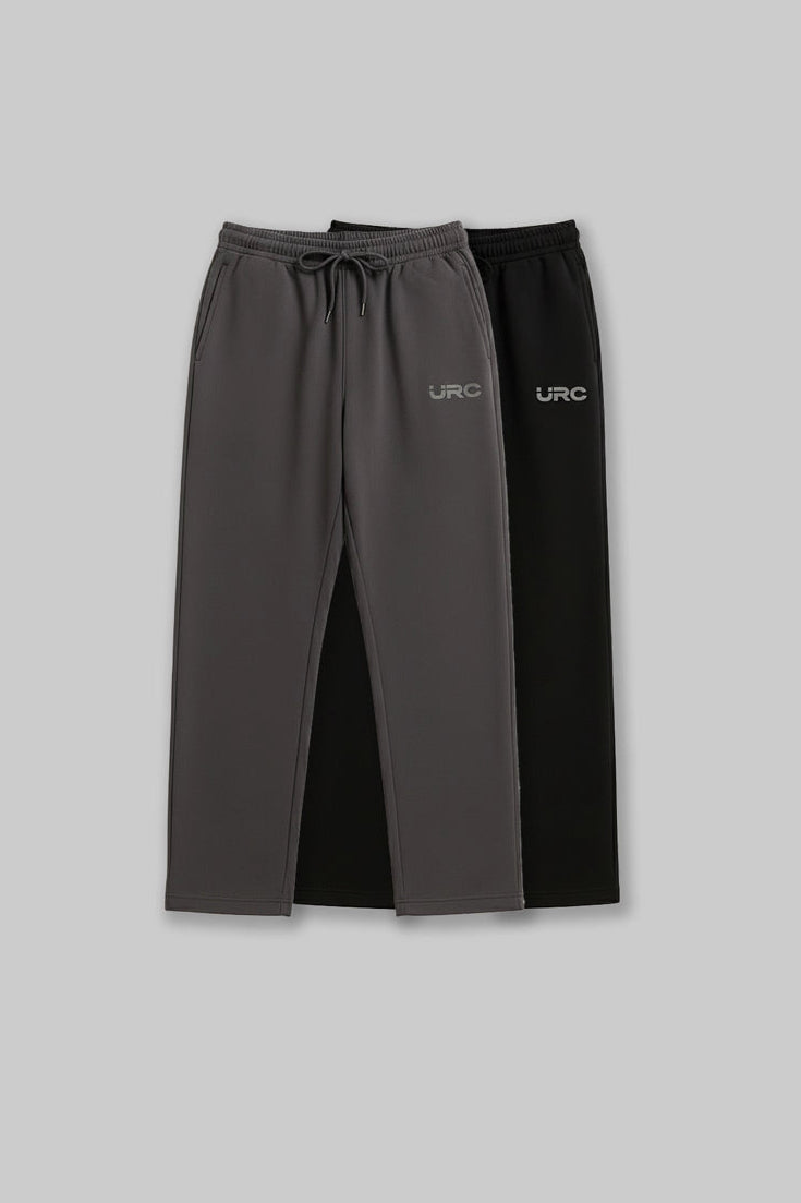 Flat lay of two pairs of relaxed-fit "URC" drawstring trousers in beige and charcoal grey.
