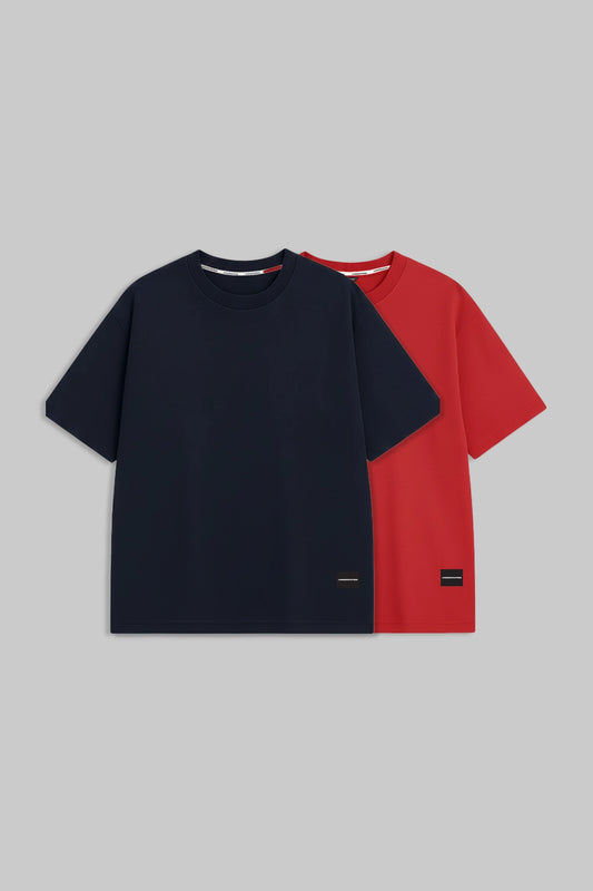 Two short-sleeve crew-neck t-shirts displayed flat against a solid gray background, with one in navy blue and the other in red. The navy shirt is positioned in front, and a small black rectangular brand patch is visible near the bottom hem.
