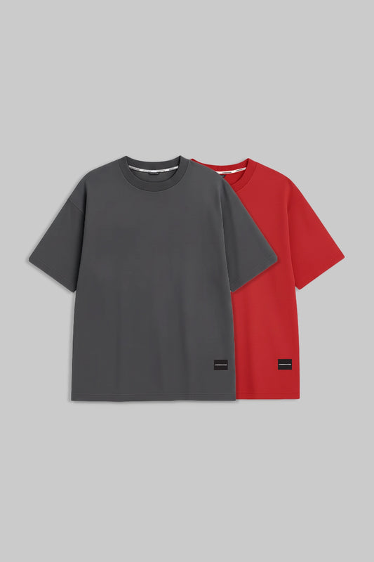 Two short-sleeve crewneck t-shirts displayed flat against a solid gray background, with one shirt in dark gray in front and one in bright red behind it. Both shirts feature a small black rectangular patch near the lower hem.
