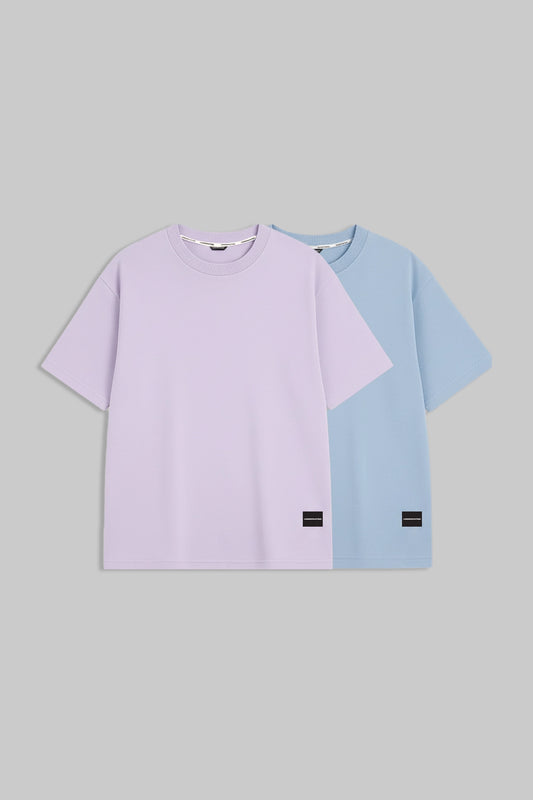 Two short-sleeve crewneck t-shirts displayed flat against a solid gray background. The front t-shirt is a light lavender purple, and the t-shirt behind it is a light blue.