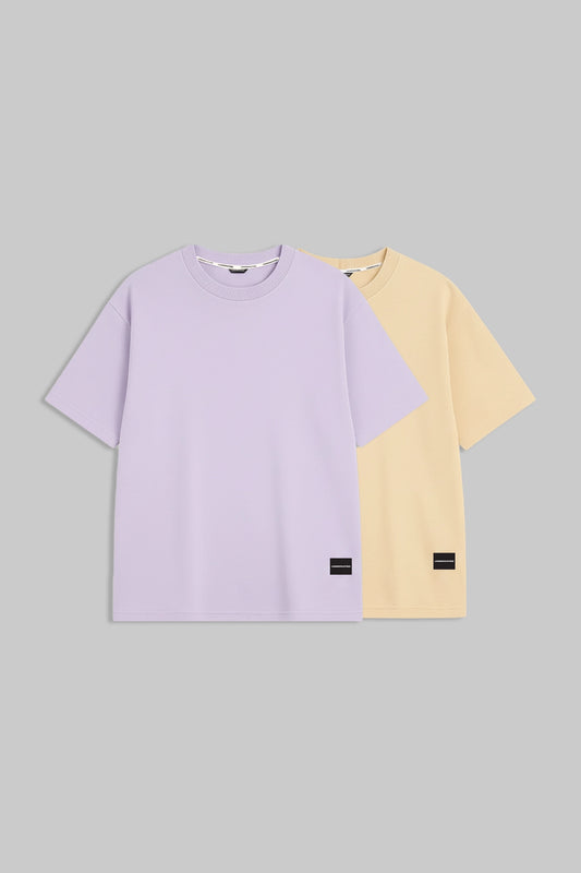 Two short-sleeve crewneck t-shirts displayed flat against a solid gray background. The front t-shirt is light lavender purple, and the t-shirt behind it is pale yellow.