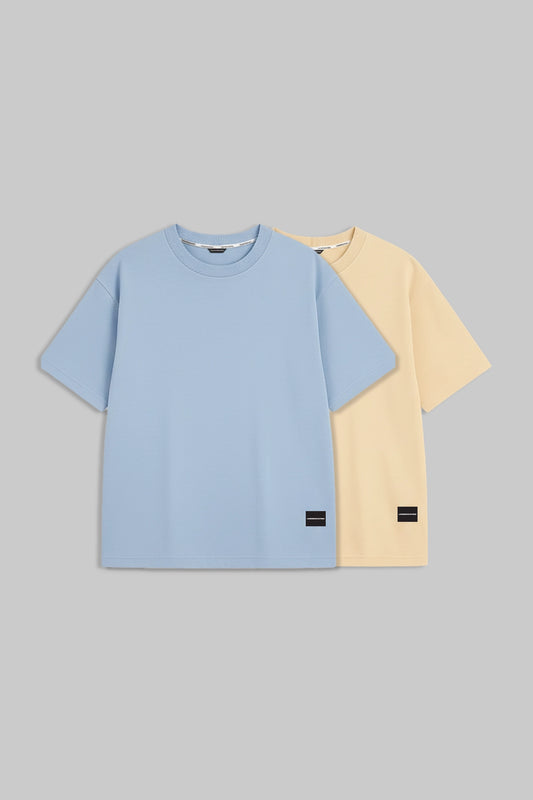 Two short-sleeve crewneck t-shirts displayed flat against a solid gray background. The front t-shirt is light blue, and the t-shirt behind it is pale yellow.