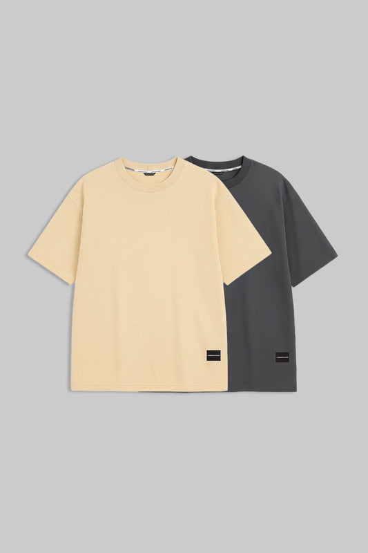 Two short-sleeve crew-neck t-shirts displayed flat against a solid gray background. The front t-shirt is a light cream/beige color, and the t-shirt behind it is dark charcoal gray.