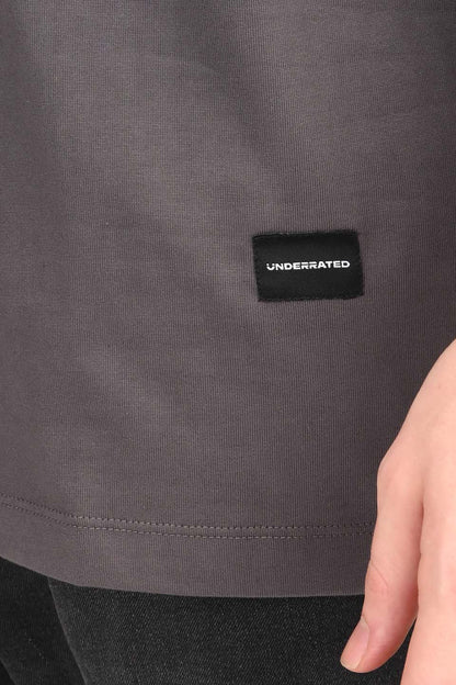 A close-up shot of the bottom right corner of a charcoal gray oversized t-shirt, showing a small black woven tag with the word 