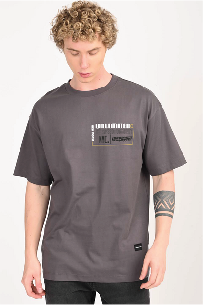 A young man with curly blonde hair wearing a charcoal gray oversized t-shirt with a rectangular graphic on the chest that includes the words 