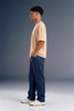 A man in profile wearing a light peach t-shirt, dark blue jeans, and white sneakers, standing against a dark gray studio background.