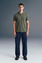 Full-length front view of a man standing straight and facing forward against a grey studio backdrop, wearing a dark olive green polo shirt and dark navy blue trousers.