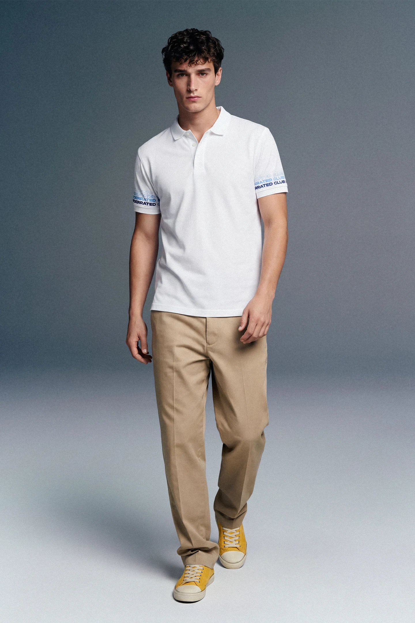 Full-length front view of a man walking forward, wearing a white polo shirt with blue-patterned cuffs, beige trousers, and yellow shoes against a grey gradient studio background.