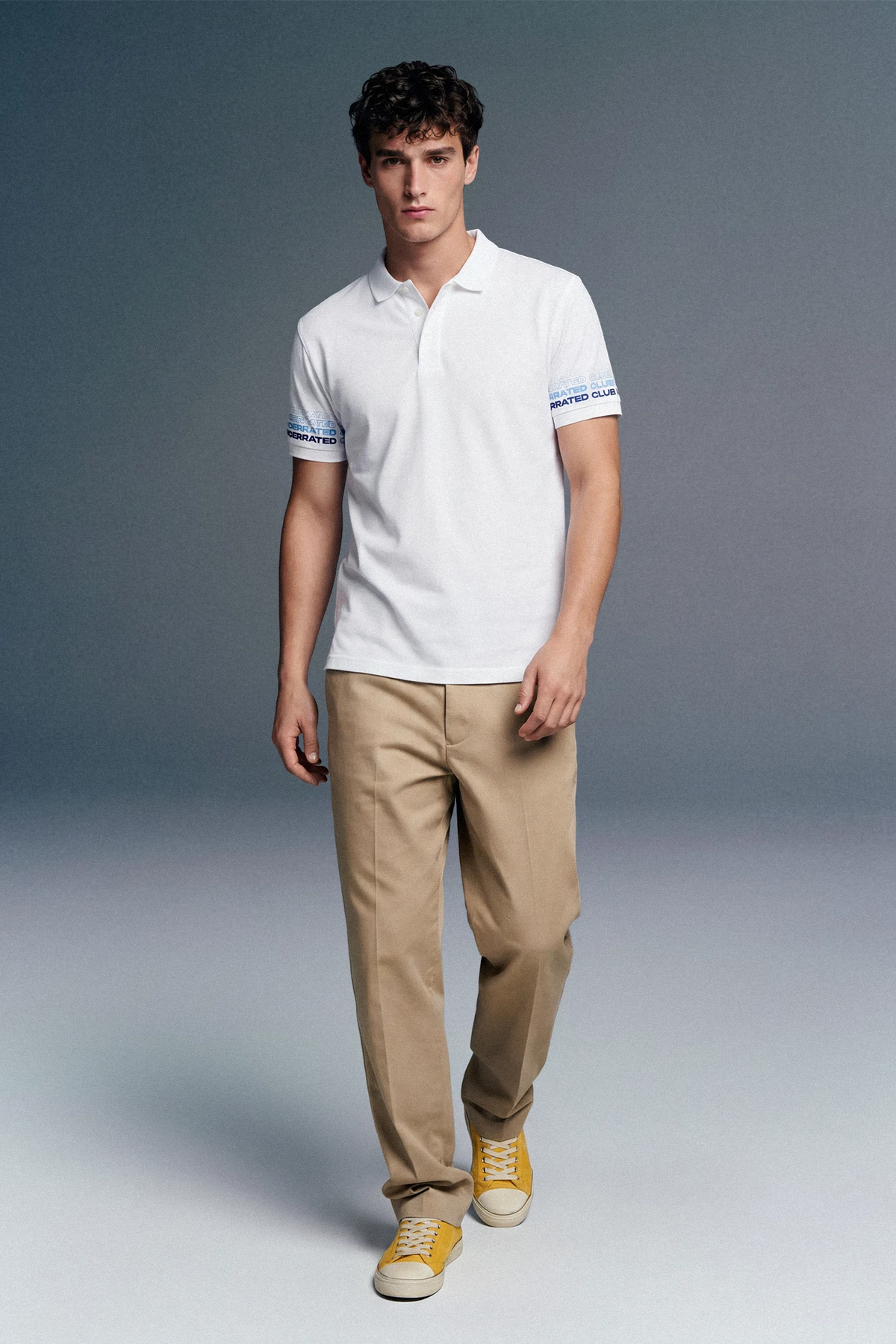 Man wearing a white polo shirt and beige pants against a gray background