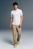 Full-length front view of a man walking forward, wearing a white polo shirt with blue-patterned cuffs, beige trousers, and yellow shoes against a grey gradient studio background.